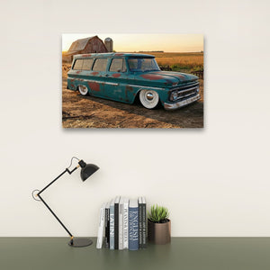 A framed wall art print showing a vintage teal 1966 Chevy Suburban Carryall parked in a rural field at sunset, displayed above a desk with a black lamp, books, and a small succulent in a neutral interior setting.