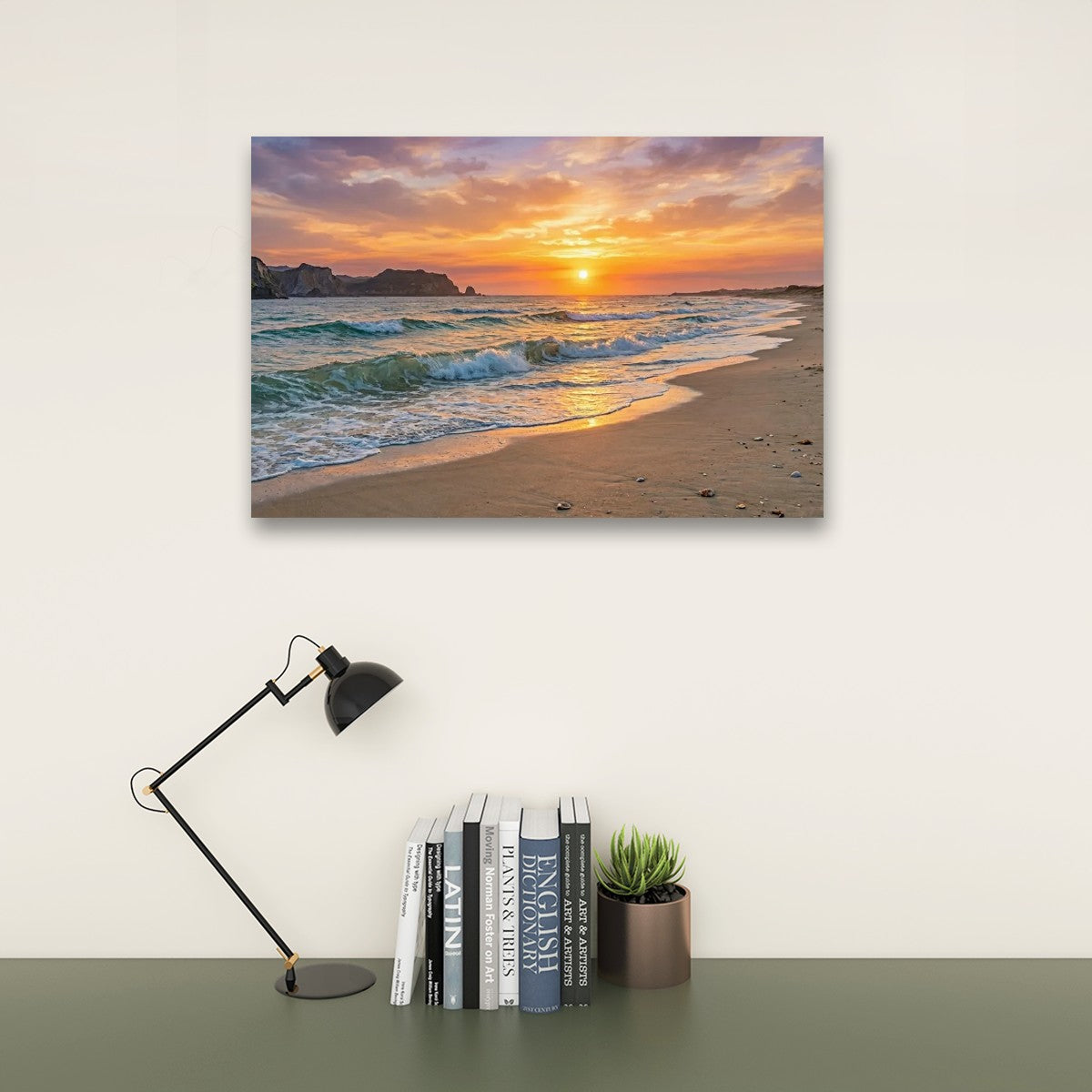 A rectangular wall art print showing a beach sunset with orange and purple sky, waves, and sand, displayed above a desk with a black lamp, books, and a small potted succulent against a light wall.