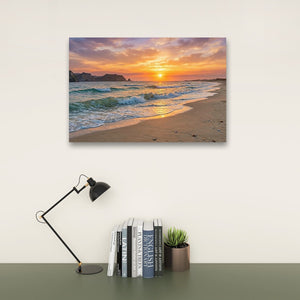 A rectangular wall art print showing a beach sunset with orange and purple sky, waves, and sand, displayed above a desk with a black lamp, books, and a small potted succulent against a light wall.