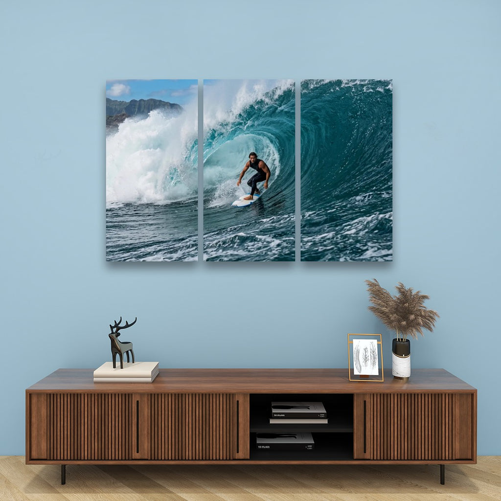 A three-panel canvas wall art print depicting a surfer inside a curling ocean wave, displayed on a light blue wall above a wooden console table with decorative items.
