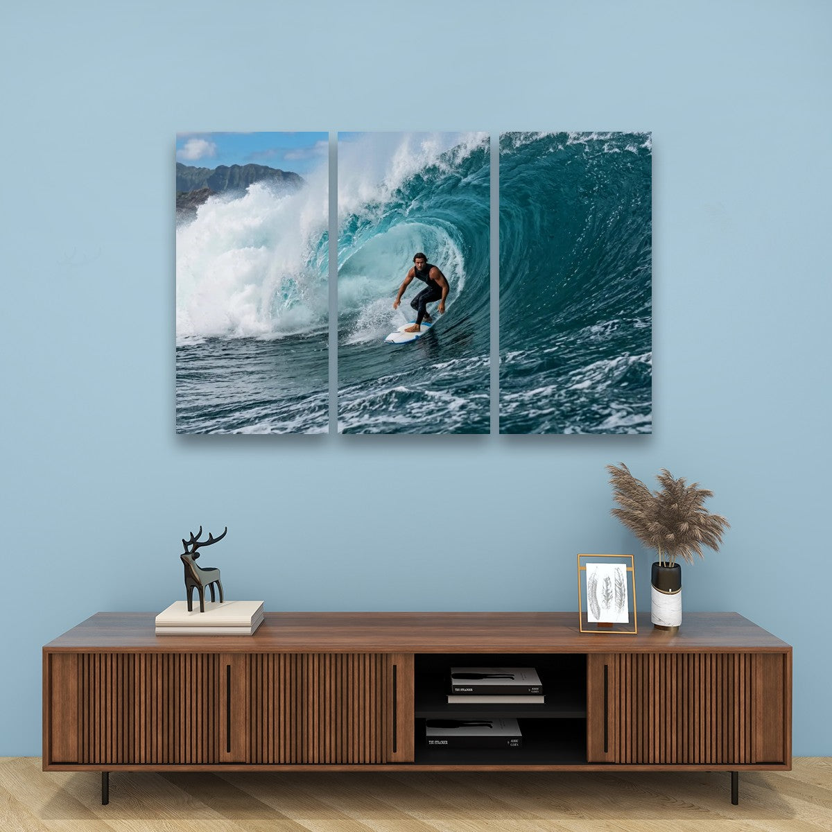 A three-panel canvas wall art print depicting a surfer inside a curling ocean wave, displayed on a light blue wall above a wooden console table with decorative items.