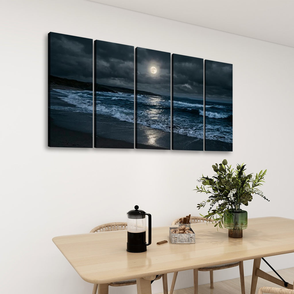 A five-panel canvas wall art set depicting a dramatic night beach scene with waves, dark clouds, and a bright full moon, displayed on a white wall above a light wood dining table with chairs and a plant.