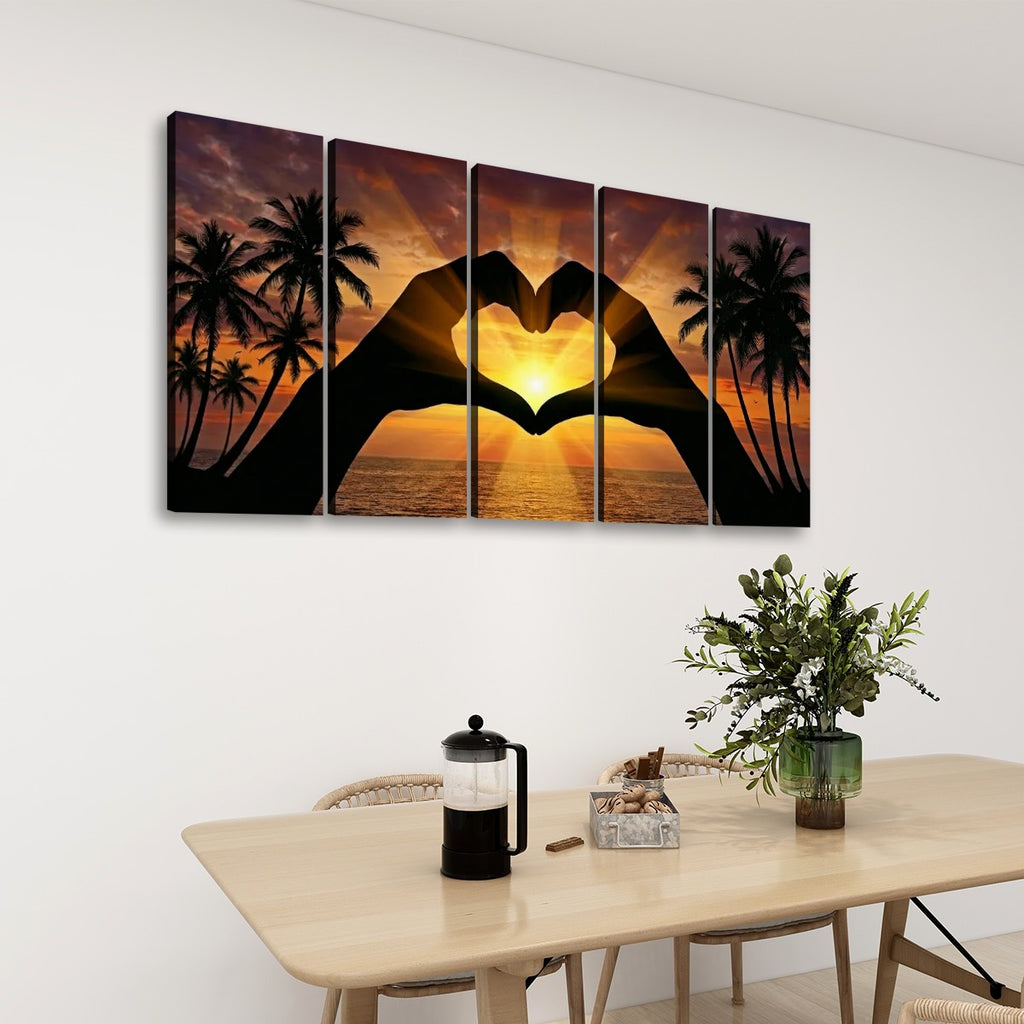 A five-panel canvas wall art set hangs above a dining table in a real interior setting. The artwork shows silhouetted hands forming a heart shape over a tropical sunset with palm trees and ocean.