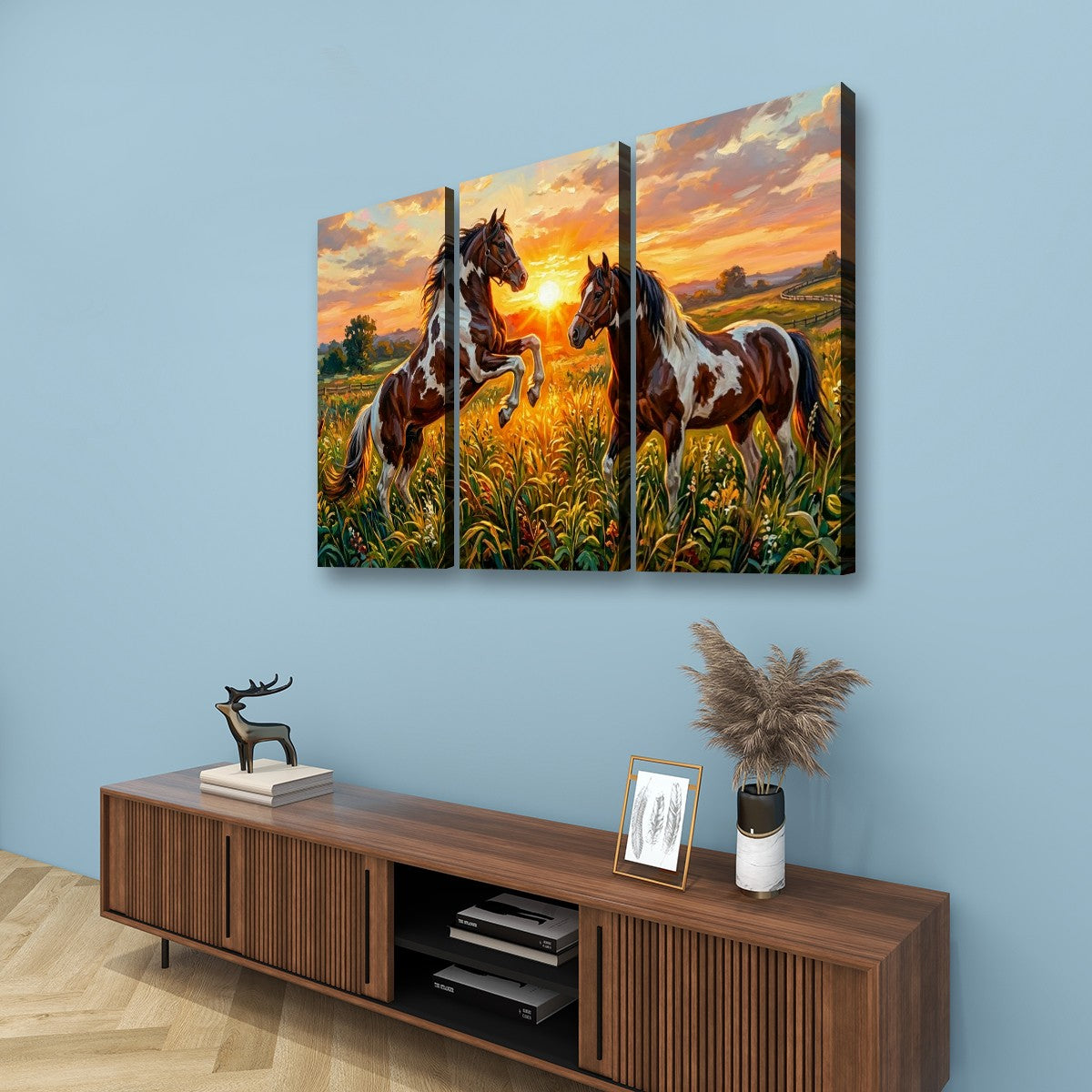 A three-panel canvas wall art set depicting two horses rearing in a golden field at sunset, displayed on a light blue wall above a wooden console table with decorative items.