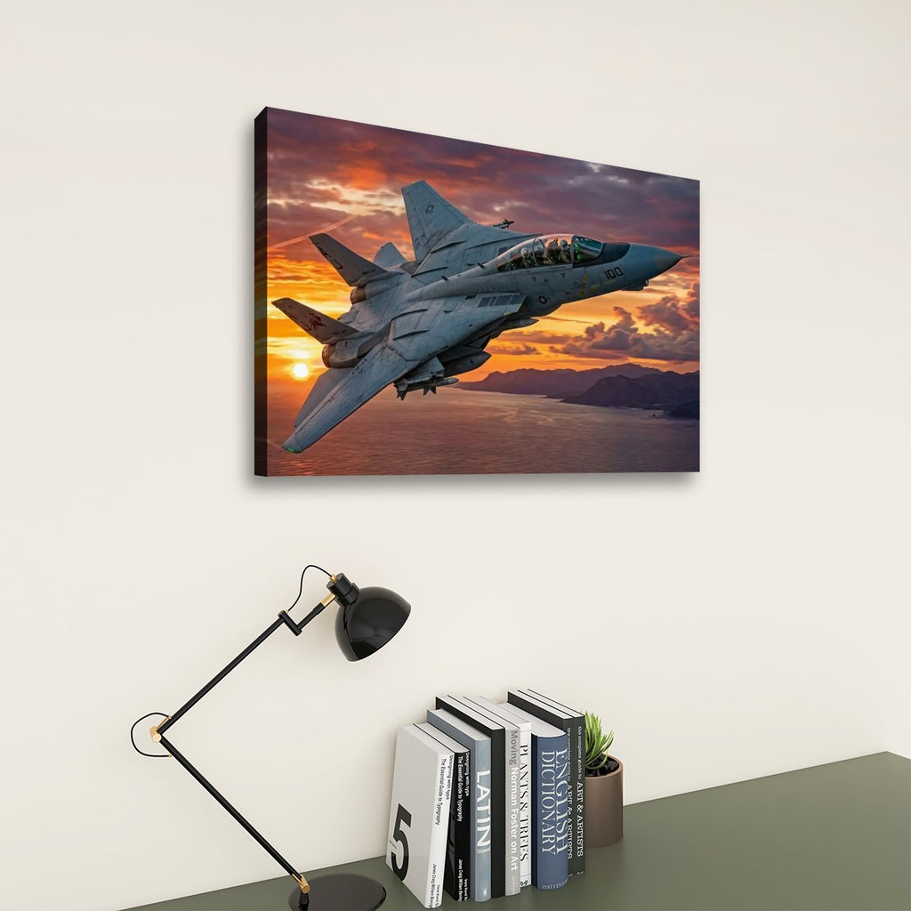 A rectangular wall art print showing a gray Grumman F-14 Tomcat  military fighter jet flying over water at sunset, displayed on a light wall above a desk with a black lamp and books.