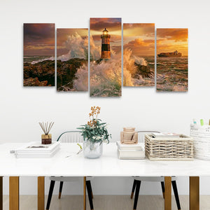 A five-panel canvas wall art set depicting a dramatic lighthouse scene at sunset with crashing waves, displayed above a white table in a modern room setting.