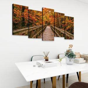A five-panel canvas wall art set depicting a wooden boardwalk bridge over autumn-colored trees, displayed on a white wall above a modern white dining table with books and a vase.