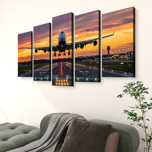A 5-panel canvas wall art set depicting an airplane landing on a runway at sunset, displayed on a white wall above a green sofa in a living room setting.