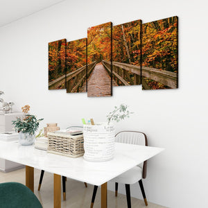 A five-panel canvas wall art set depicting a wooden boardwalk bridge over autumn foliage, displayed on a white wall above a dining table in a modern room setting.