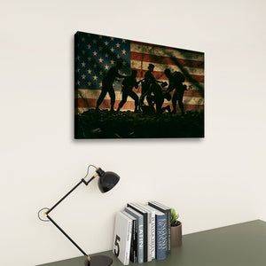 A rectangular wall art print hanging on a light wall above a desk, showing silhouetted figures in front of a distressed American flag. The scene includes a black desk lamp, stacked books, and a small plant below the artwork.
