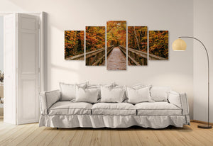 A 5-panel canvas wall art set depicting an autumn forest pathway over a wooden bridge, displayed on a white wall above a light gray sofa in a modern living room setting.