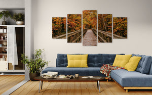 A 5-panel canvas wall art set depicting an autumn forest pathway over a wooden bridge, displayed on a wall above a blue sectional sofa in a modern living room setting.