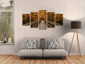 A 5-panel canvas wall art set depicting an autumn forest pathway over a wooden bridge, displayed on a light gray wall above a modern gray sofa in a living room setting.