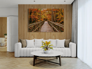 A three-panel canvas wall art print depicting a wooden boardwalk bridge over autumn foliage, displayed on a wood-paneled wall above a modern white sofa in a bright living room.