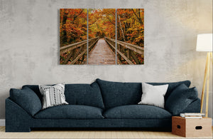 A three-panel canvas wall art print depicting a wooden boardwalk bridge over autumn foliage in a forest, displayed on a textured gray wall above a dark blue sofa in a living room setting.