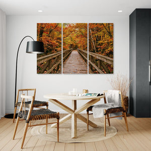 A three-panel canvas wall art print depicting a wooden boardwalk bridge over autumn foliage, displayed in a modern dining room setting above a light wood table and chairs.