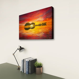A rectangular wall art print or canvas showing a colorful sunset over water with a guitar-shaped silhouette and reflections, displayed on a light wall above a desk with a lamp, books, and a small plant.