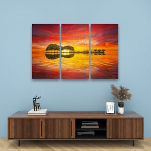 A three-panel canvas wall art set depicting a vibrant sunset over a lake with tree silhouettes and reflections, displayed on a light blue wall above a wooden console table with decorative items.