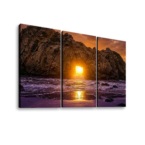 Three-panel canvas wall art showing a dramatic rocky coastline at sunset, with the sun low over the ocean and purple-toned waves reflecting on the shore, displayed against a white background.