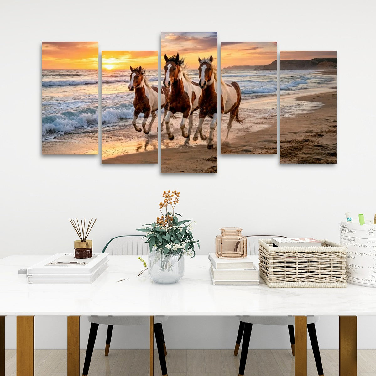 A five-panel canvas wall art set depicting three horses running along a beach at sunset, displayed on a white wall above a styled white table with books, a vase, and baskets.