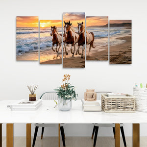 A five-panel canvas wall art set depicting three horses running along a beach at sunset, displayed on a white wall above a styled white table with books, a vase, and baskets.
