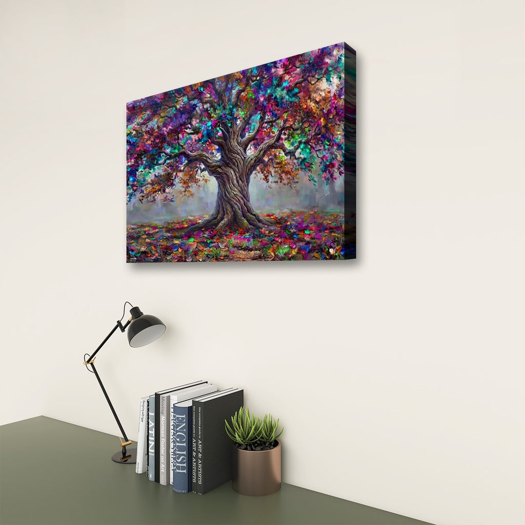A colorful canvas wall art print depicting a stylized tree with vibrant multicolored foliage, shown hanging on a light beige wall above a desk with a lamp, books, and a small plant.