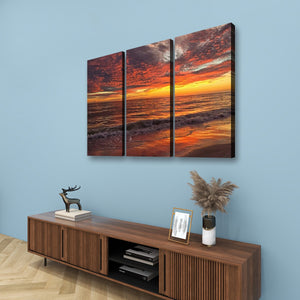Three-panel canvas wall art showing a dramatic sunset over the ocean, displayed above a wooden console table against a light blue wall.