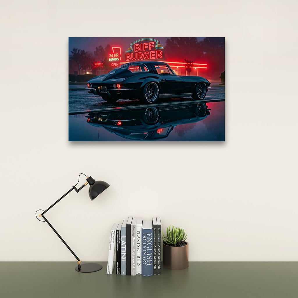 A framed wall art print showing a dark 1963 Split Window Chevy Corvette at night with a glowing red 'Biff Burger' neon sign and its reflection in wet pavement, displayed above a desk with a black lamp, books, and a small succulent.