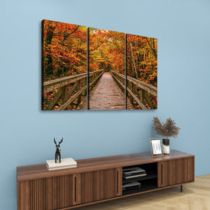 A three-panel canvas wall art set depicting a wooden boardwalk bridge over autumn foliage in orange and red tones, displayed on a light blue wall above a wooden console table.