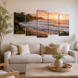 A five-panel canvas wall art set depicting a coastal sunset beach scene, displayed above a beige sofa in a modern, neutral-toned living room.