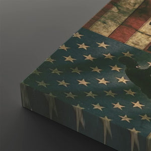 A close-up of a rectangular canvas or print featuring a distressed American flag design, with a dark silhouette shape over the stars, photographed on a dark gray background.