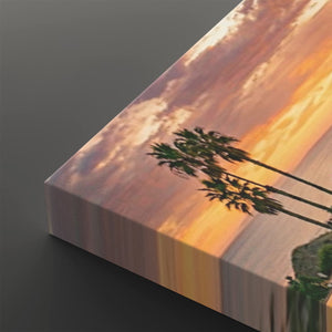A rectangular canvas wall art print shown at an angle on a dark gray background, featuring a sunset scene with palm tree silhouettes against orange, pink, and purple sky tones.