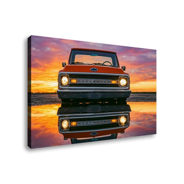 A rectangular wall art print or canvas showing a front-facing vintage orange  1970 Chevrolet C10 truck parked on reflective water at sunset, with a dramatic pink, orange, and purple sky. The print is photographed at a slight angle against a plain white background.