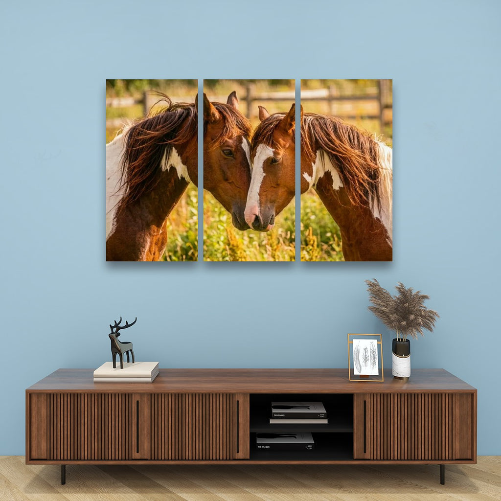A three-panel canvas wall art print depicting two brown horses with white markings facing each other in a sunlit field, displayed on a light blue wall above a wooden console table with decorative items.