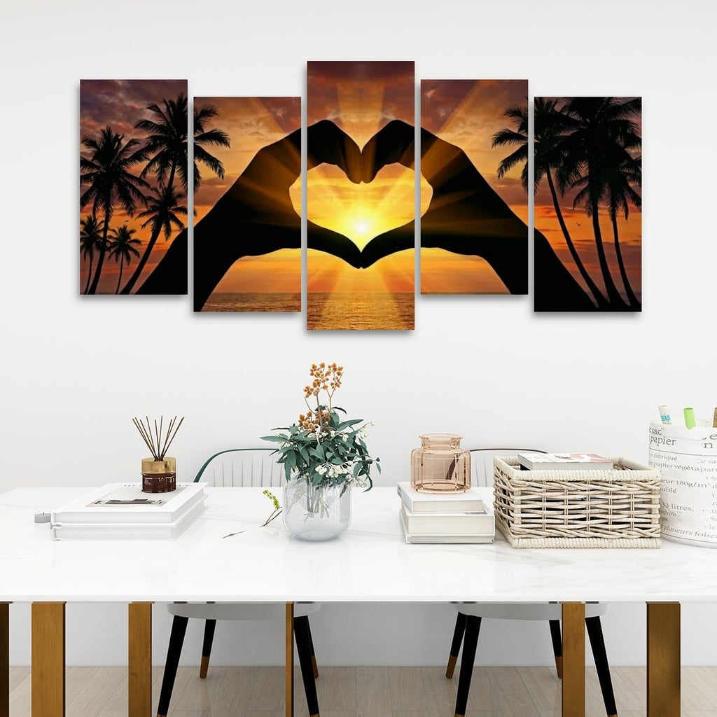A five-panel canvas wall art set depicting a sunset over a tropical beach with palm trees and two silhouetted hands forming a heart shape, displayed above a white dining table in a modern room setting.