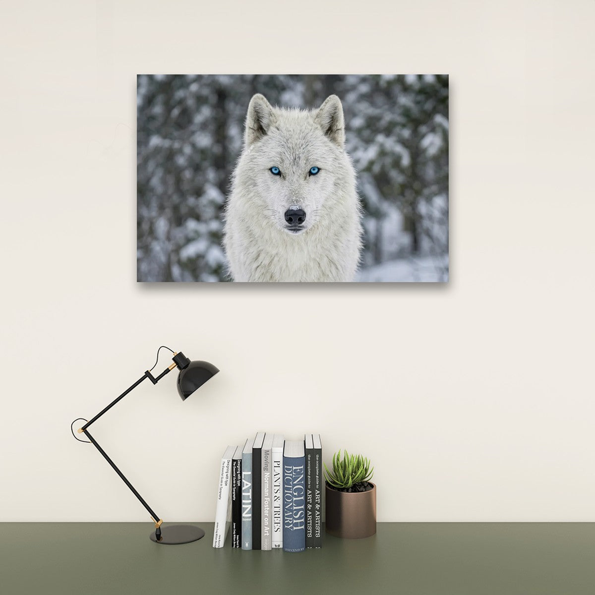 A rectangular wall art print showing a white wolf with bright blue eyes in a snowy outdoor setting, displayed above a desk with a black lamp, books, and a small succulent against a beige wall.
