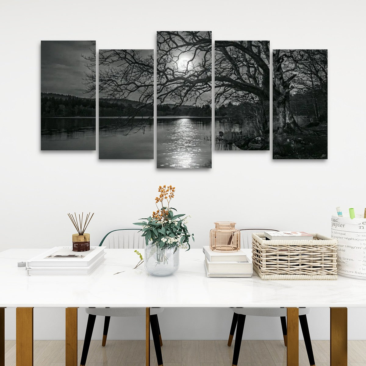 A five-panel black and white canvas wall art set depicting a moody lakeside scene with bare trees and moonlit reflections, displayed above a white dining table in a modern room setting.