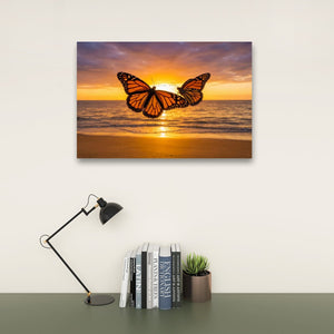 A rectangular wall art print showing a monarch butterfly over a beach sunset, displayed above a desk with books, a small succulent, and a black desk lamp against a light wall.