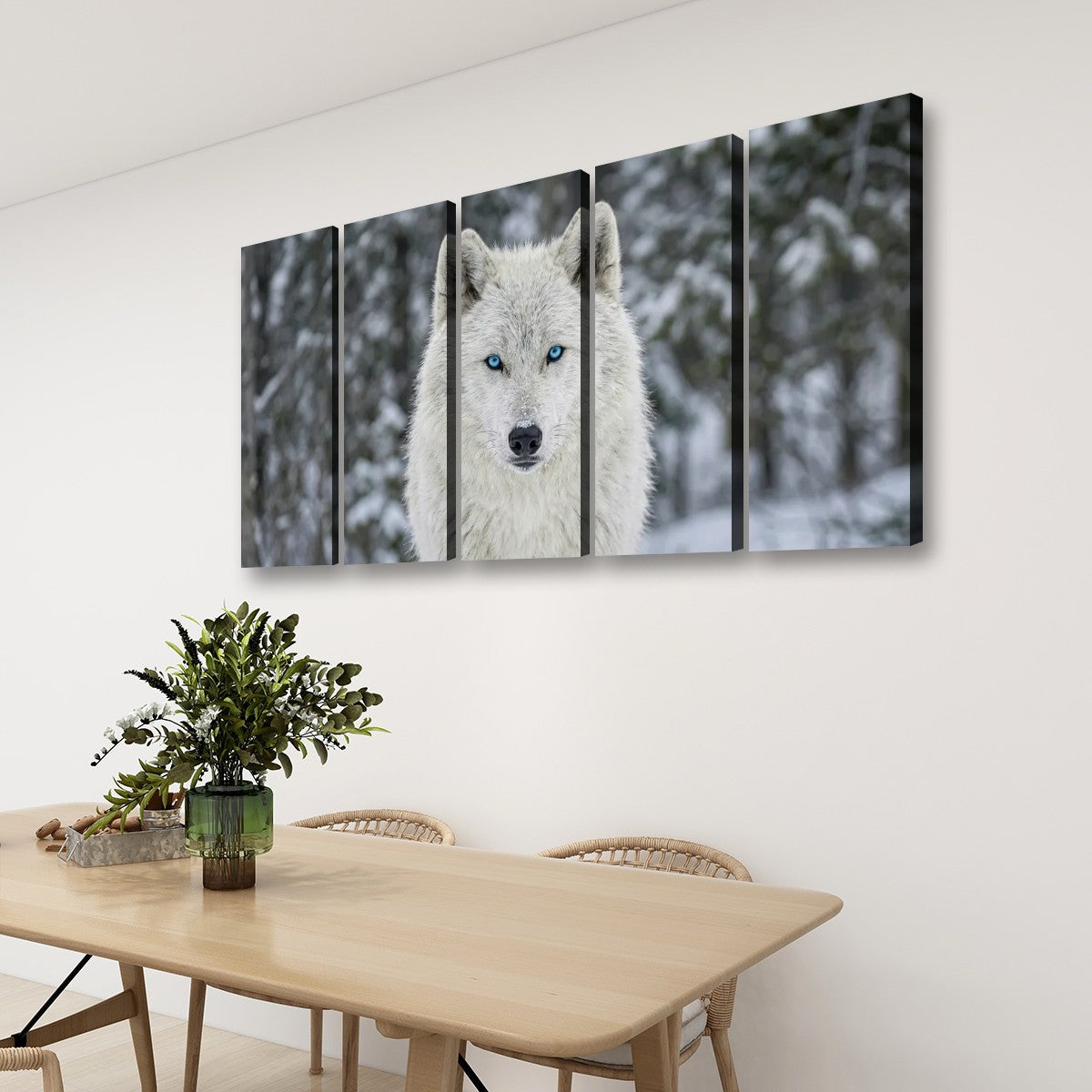 A four-panel canvas wall art set depicting a close-up portrait of a white wolf with blue eyes in a snowy forest, displayed on a white wall above a wooden dining table with chairs and a plant.