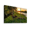 Three-panel canvas wall art showing a family of ducks standing in grass beside a lake at sunset, with trees and water in the background.