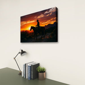 A rectangular wall art print or canvas showing a silhouetted rider on a horse against a vivid sunset sky, displayed on a light wall above a desk with a lamp, books, and a small plant.