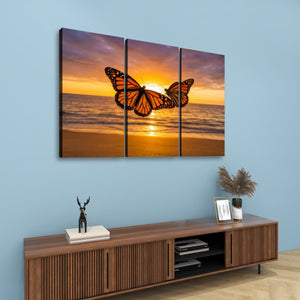 A three-panel canvas wall art set depicting a monarch butterfly over a golden sunset beach, displayed on a light blue wall above a wooden console table with decorative items.