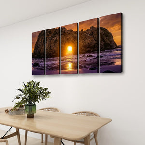 A four-panel canvas wall art set depicting a dramatic rocky coastline at sunset, displayed on a white wall above a light wood dining table with chairs and a green plant.