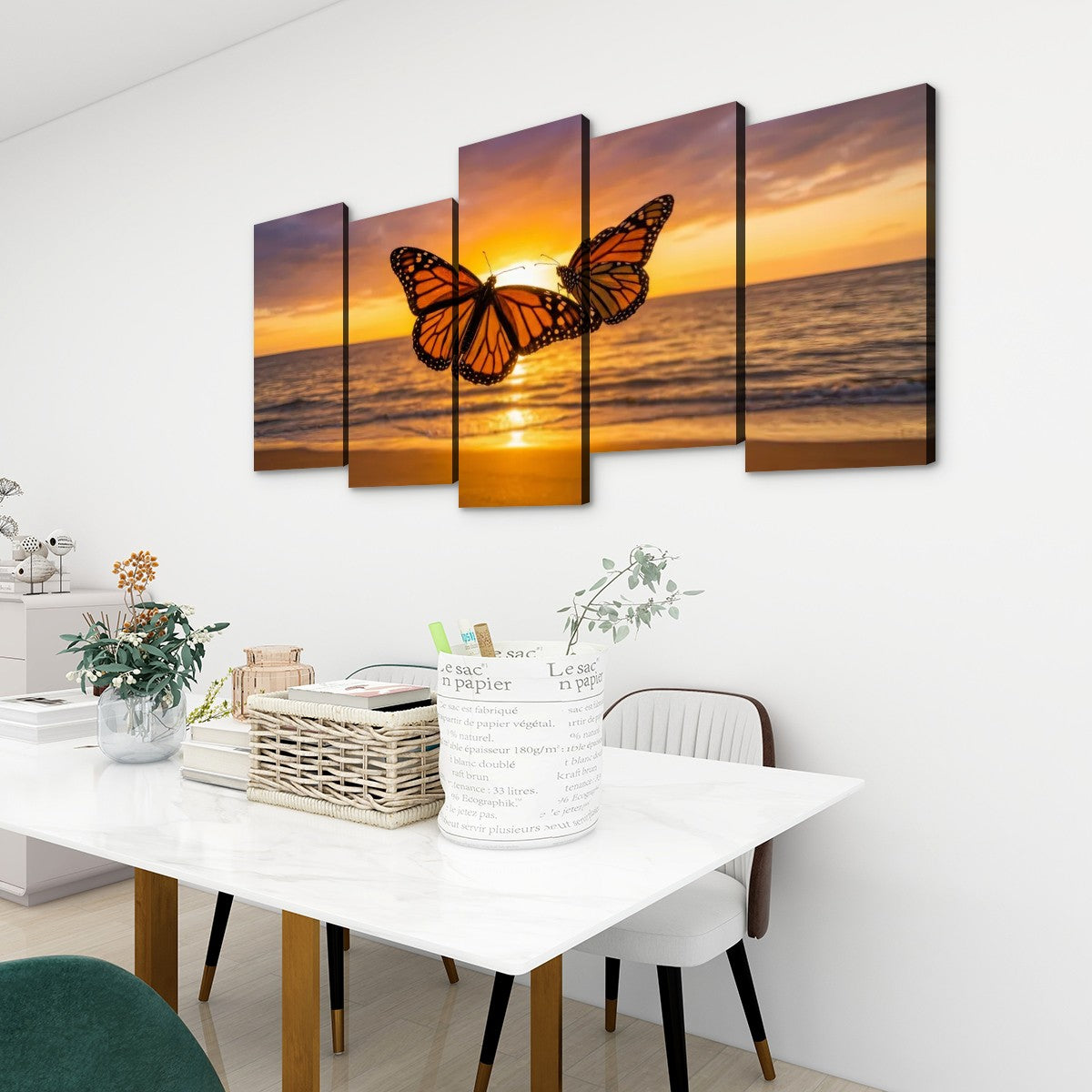 A 5-panel canvas wall art set depicting a monarch butterfly over a golden sunset ocean, displayed on a white wall above a dining table in a modern room setting.