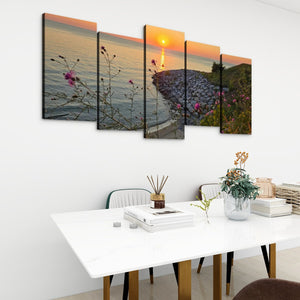 A five-panel canvas wall art set depicting a coastal sunset over water with flowers and a rocky shoreline, displayed above a white dining table in a modern room setting.