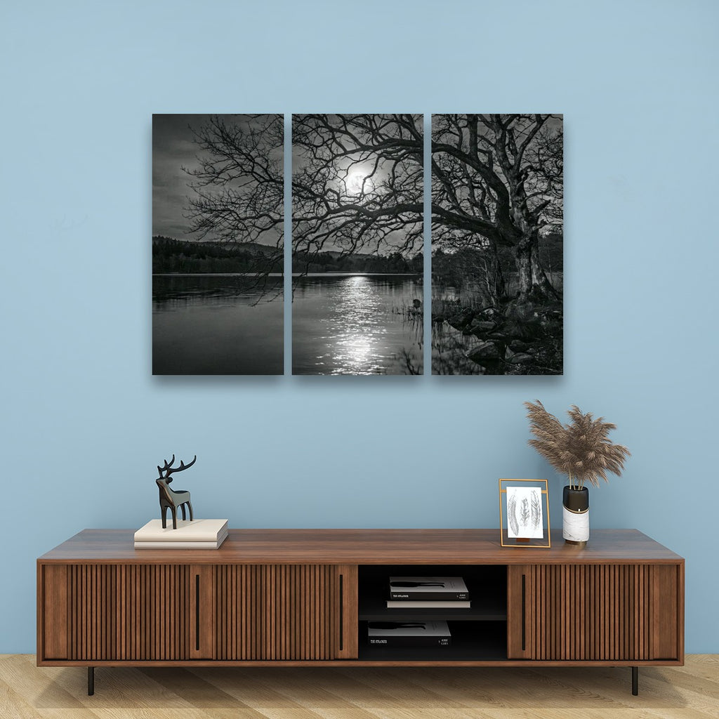 A three-panel black and white canvas wall art set depicting a moonlit lake scene with bare trees, displayed above a wooden console cabinet in a styled room with a light blue wall.