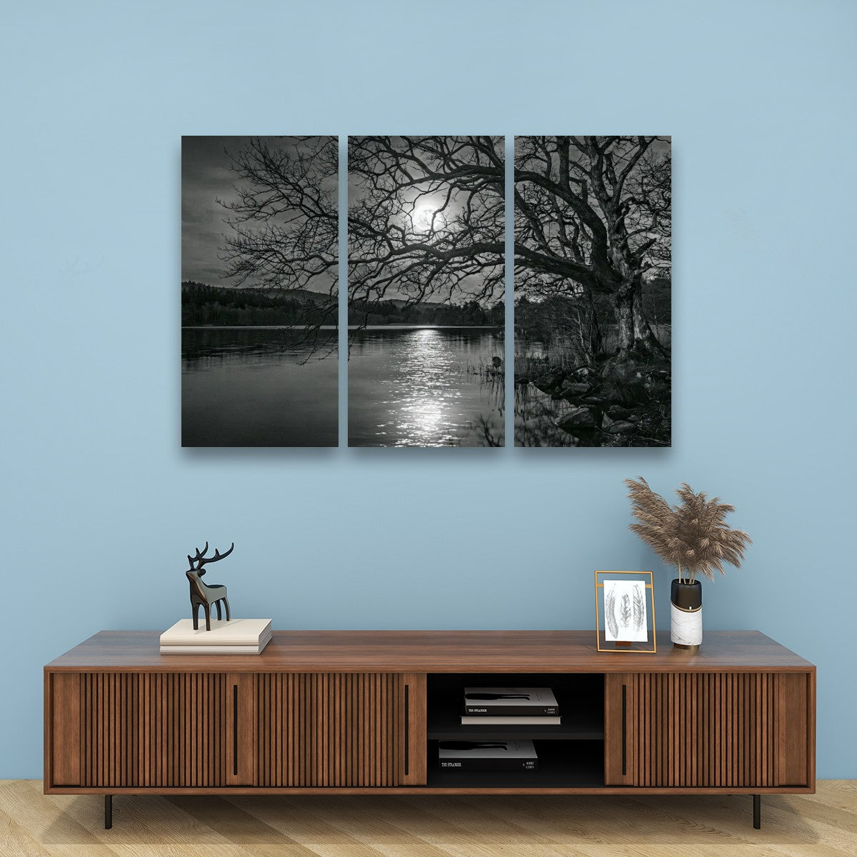 A three-panel black and white canvas wall art set depicting a moonlit lake scene with bare trees, displayed above a wooden console cabinet in a styled room with a light blue wall.