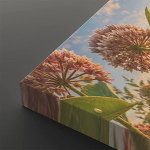 A square canvas wall art print shown at an angle, featuring a close-up floral scene with pink flower clusters, green leaves, and a blue sky background.