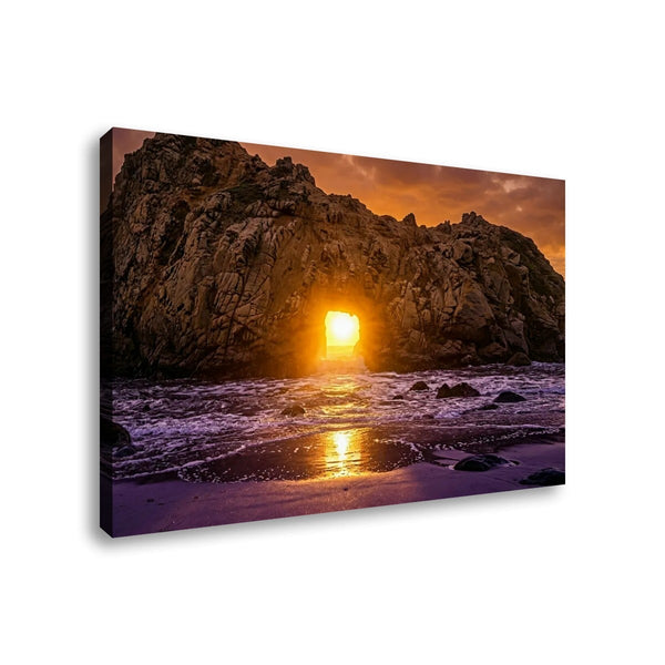 A rectangular canvas wall art print showing a rocky beach at sunset, with the sun shining through the rocks and reflecting on the water. The canvas is photographed at a slight angle against a plain white background.
