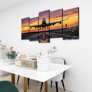 A 5-panel canvas wall art set depicting a commercial airplane landing during sunset over an airport runway, displayed on a white wall above a dining table in a modern room setting.